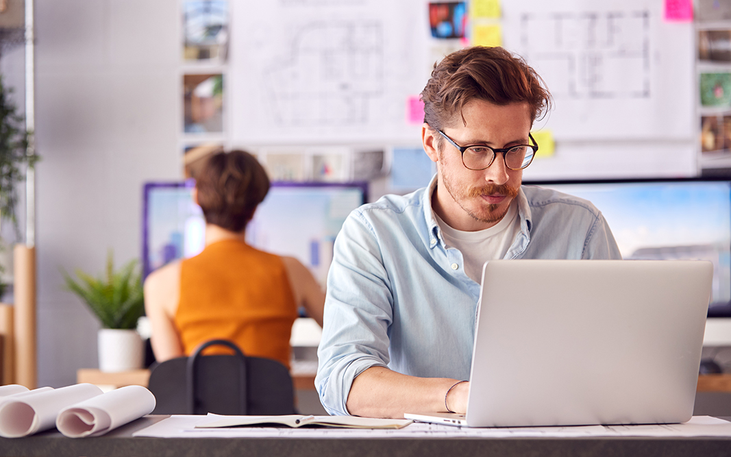 Male And Female Architects In Office Working At Desk On Laptop And Desktop Computers